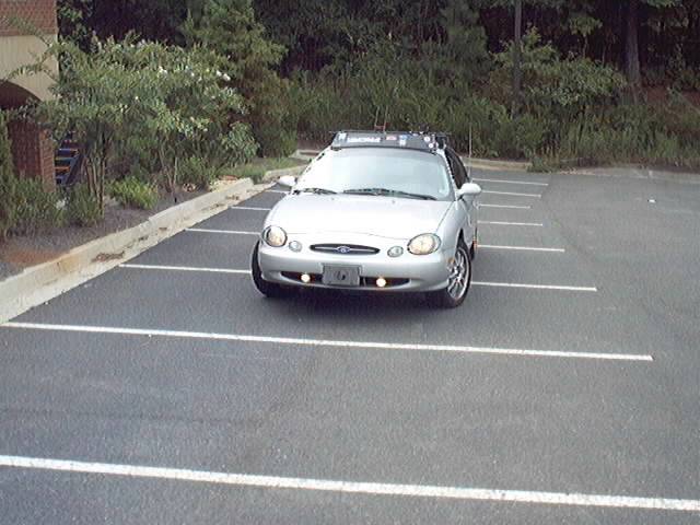 Silver Ford Taurus sedan front view in an empty parking lot with fog lights and roof crossbars