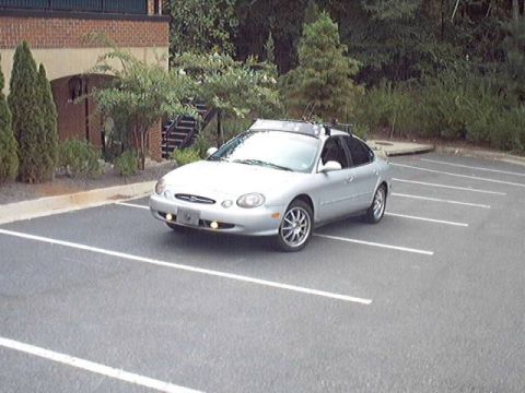 1998 Ford Taurus - A silver Ford Taurus sedan parked at an angle in an empty striped asphalt lot beside a brick building and landscaped shrubs, shot from the…