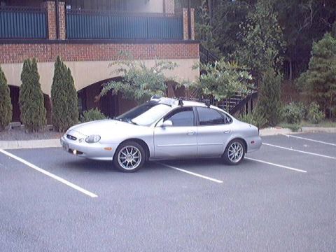 1998 Ford Taurus - A silver Ford Taurus four-door sedan parked in an empty striped lot, shot from the front three-quarter side. It wears aftermarket multi-spo…