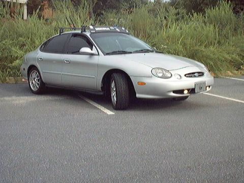 1998 Ford Taurus - Silver four-door Ford Taurus sedan photographed from the front three-quarter angle in an empty asphalt parking lot backed by tall grass and…