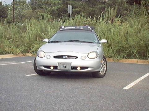 1998 Ford Taurus - Front view of a silver Ford Taurus sedan parked in an empty asphalt lot beside tall grass, with round fog lights lit in the lower bumper an…