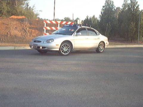 1998 Ford Taurus - A silver Ford Taurus sedan with chrome mesh-style aftermarket wheels and a roof rack sits on fresh asphalt, viewed from the front three-qua…