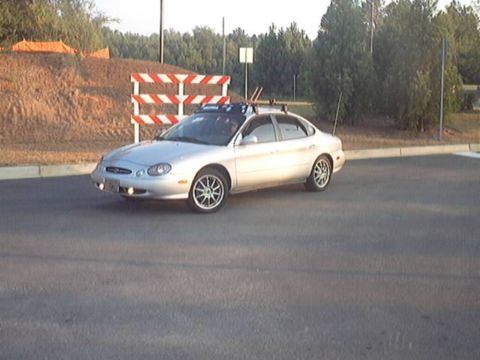 1998 Ford Taurus - Three-quarter front view of a silver Ford Taurus sedan parked on fresh asphalt, fitted with a roof rack, aftermarket multi-spoke alloy whee…