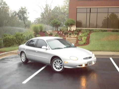 1998 Ford Taurus - A silver late-1990s Ford Taurus sedan parked at an angle across the stalls of a wet parking lot, shown from the front three-quarter with it…