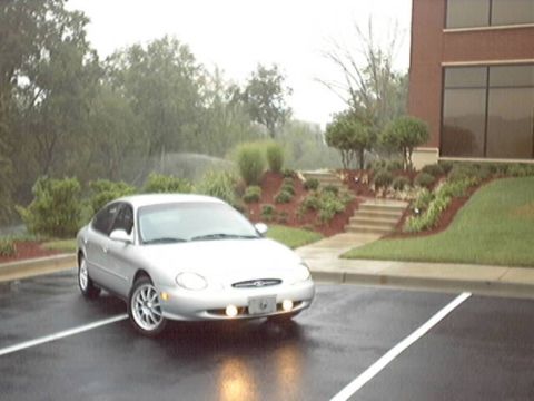 1998 Ford Taurus - Silver Ford Taurus sedan shown front-three-quarter in a wet parking lot with its fog lights glowing, beside a brick office building with ti…