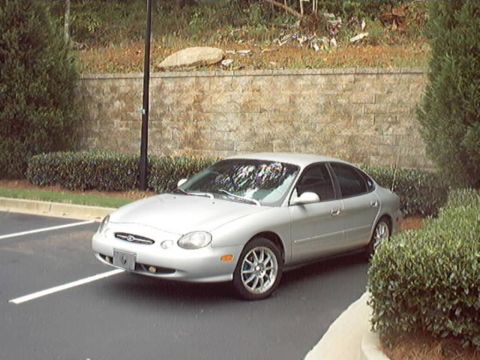 1998 Ford Taurus - A silver Ford Taurus sedan parked in a striped lot, shot from an elevated front three-quarter angle showing the oval grille, projector-styl…