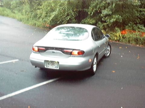 1998 Ford Taurus - Silver Ford Taurus LX sedan sitting on damp asphalt, shot from an elevated three-quarter rear angle that shows the full-width oval tail lam…