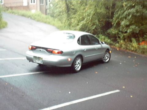 1998 Ford Taurus - A silver sedan is parked on a wet asphalt surface, with trees and a building in the background.