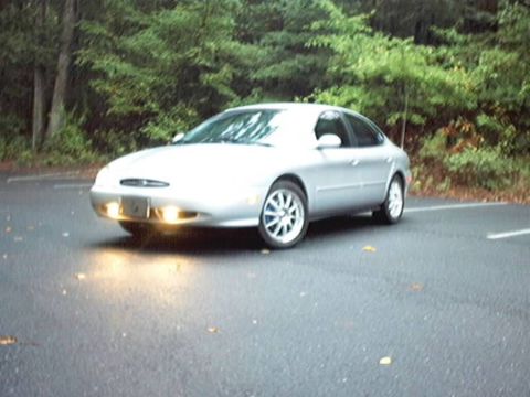 1998 Ford Taurus - A silver late-1990s Ford Taurus sedan angled on damp leaf-strewn pavement in front of dense green woods, front three-quarter view with its…