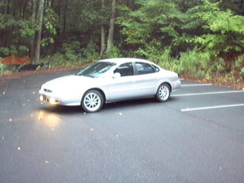 1998 Ford Taurus - Silver four-door Ford Taurus sedan photographed from the driver's side in a wet asphalt parking lot backed by dense green woods. The car ha…