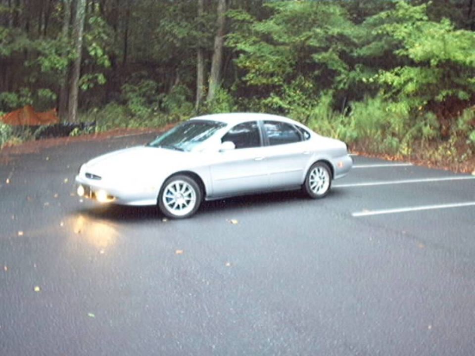 Silver Ford Taurus sedan side view in wet parking lot with headlights on, woods behind