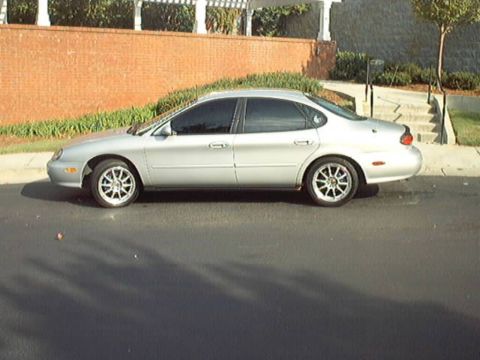 1998 Ford Taurus - Full side profile of a silver Ford Taurus sedan parked at a curb, showing tinted windows, chrome multi-spoke aftermarket wheels and the rou…