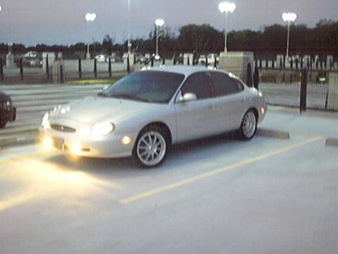 1998 Ford Taurus - A silver sedan is parked in a lot as the sky darkens, with its headlights illuminated.