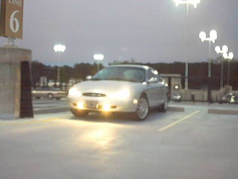 1998 Ford Taurus - A silver 1998 Ford Taurus sedan is parked in a lot as dusk settles, with its headlights on.