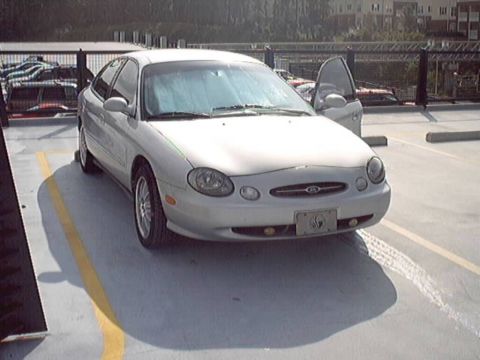 1998 Ford Taurus - A silver 1998 Ford Taurus is parked in a lot with its driver's door ajar.
