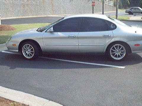 1998 Ford Taurus - Side view of a silver Ford Taurus sedan angled into a striped parking space, wearing dark tinted glass and chrome multi-spoke aftermarket w…