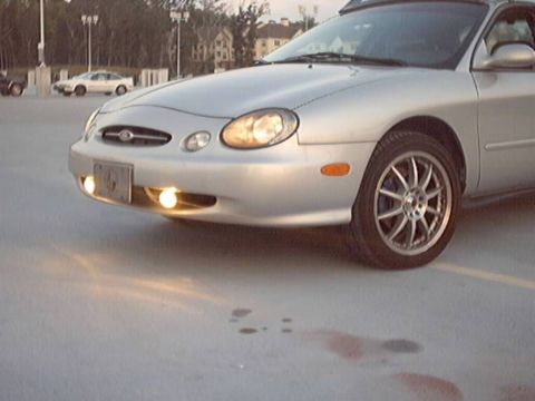 1998 Ford Taurus - A silver 1998 Ford Taurus is parked outside, its front end angled towards the camera with the headlights illuminated.