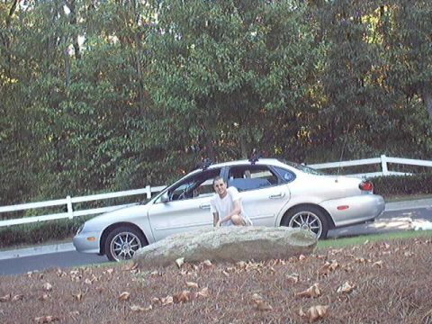 1998 Ford Taurus - A silver Ford Taurus sedan with a roof rack and chrome aftermarket wheels parked on a driveway, with a person in a white t-shirt seated on…