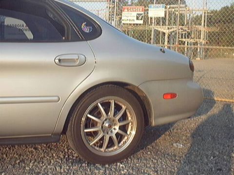 1998 Ford Taurus - Close-up of the rear quarter panel and wheel of a silver Ford Taurus sedan on gravel, showing a multi-spoke silver alloy wheel with polishe…