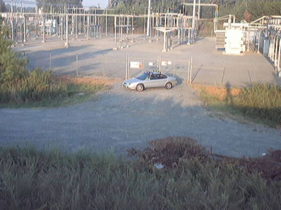 Silver sedan with roof rack parked inside fenced area of electrical substation