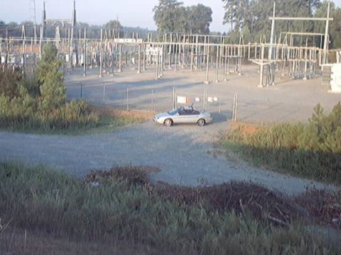 1998 Ford Taurus - Distant elevated view of a silver four-door sedan with a roof rack parked on gravel outside a chain-link-fenced electrical substation. Tall…