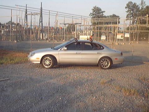 1998 Ford Taurus - A silver 1998 Ford Taurus sedan is parked on a gravel lot near an electrical substation.
