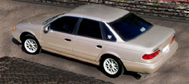 1993 Ford Taurus - Small elevated three-quarter rear view of a champagne/silver four-door sedan with an aero-rounded body, photographed on a dark paved surfac…