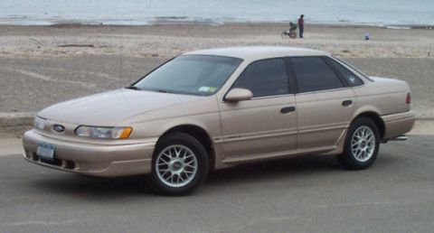 1993 Ford Taurus - Three-quarter front view of a champagne/tan Ford four-door sedan parked on pavement at a beach, with sand, surf and a person with a bicycle…