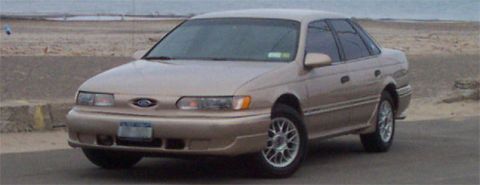 1993 Ford Taurus - Front three-quarter view of a champagne/rose 1990s Ford Taurus sedan parked on pavement near a beach, wearing a smooth body-color front bum…