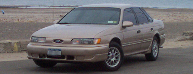 Champagne 1990s Ford Taurus sedan, front three-quarter view near a beach