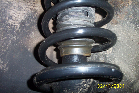 1993 Ford Taurus - Close-up of a black-painted coil spring installed over a shock/strut body, photographed from below in a dirty wheel well with road grime co…