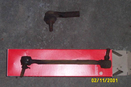 1993 Ford Taurus - A rusted, worn tie rod end lies on a concrete garage floor above a new replacement tie rod assembly resting in its open red parts box. A 02…