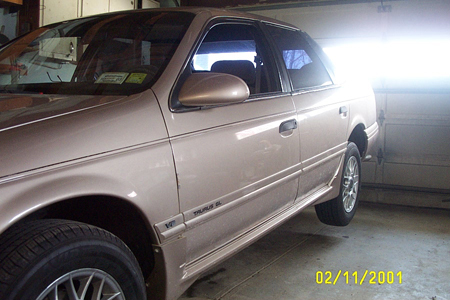 1993 Ford Taurus - Low side view of a rose/mauve four-door sedan sitting inside a garage with the door open behind it, showing the body-side molding, factory-…