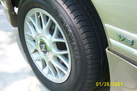 1993 Ford Taurus - Close-up of the front wheel of a light gold/champagne car fitted with a silver multi-spoke mesh alloy wheel and a Yokohama tire, with a chr…