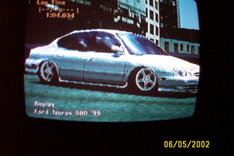 1993 Ford Taurus - Photo of a CRT television screen showing a racing video game replay of a silver Ford Taurus SHO sedan in side profile on a city street, wit…