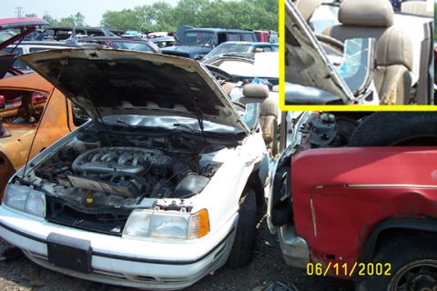 1993 Ford Taurus - Salvage-yard photo of a wrecked white sedan with the hood raised, exposing a V6 engine with intake runners, alongside rows of other junked…
