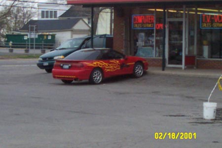 1993 Ford Taurus - A red sports car with flame decals is parked next to a dark SUV in front of a store.