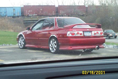 1993 Ford Taurus - A red sedan with a spoiler and custom wheels is parked near a train track with cargo containers.