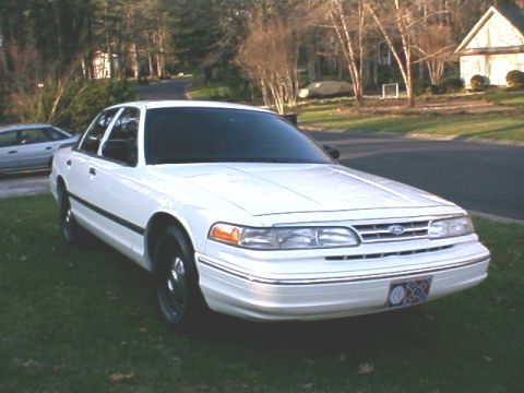 1996 Ford Crown Victoria - A white Ford Crown Victoria is parked on a grassy area next to a road, with a house and trees in the background.