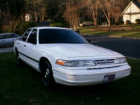 1996 Ford Crown Victoria - A white sedan is parked on a grassy area in front of a house with trees in the background.