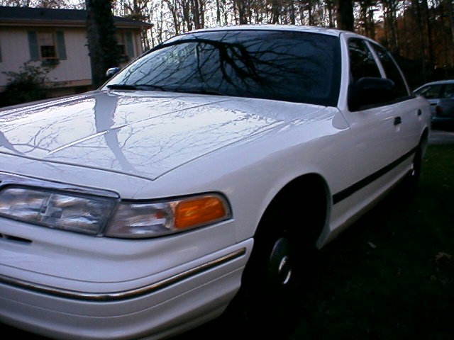 White full-size sedan, front 3/4 low angle, parked on grass by a house