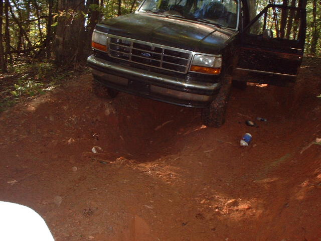 Dark Ford Bronco with open door perched above a deep red-clay hole on a wooded off-road trail