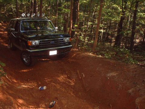 1990 Ford Bronco - A black Ford Bronco navigates a wooded trail, with two cans and a phone on the ground nearby.