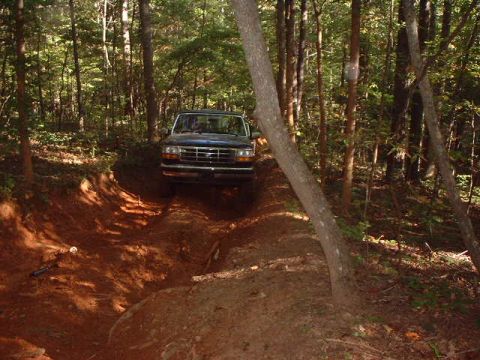 1990 Ford Bronco - A dark blue Ford Bronco navigates a wooded trail, its front grille clearly visible.