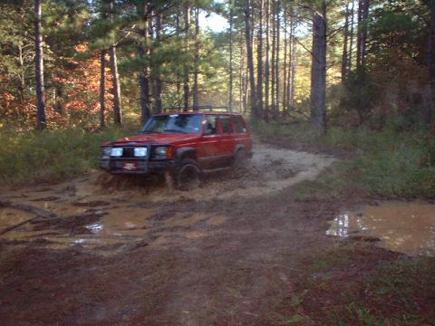 1990 Ford Bronco - A red Jeep Cherokee XJ navigates a muddy trail surrounded by trees.