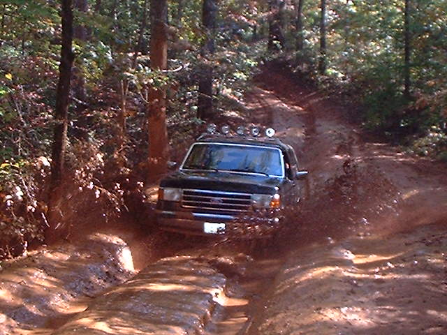 Dark Ford SUV with roof light bar driving through muddy forest trail ruts