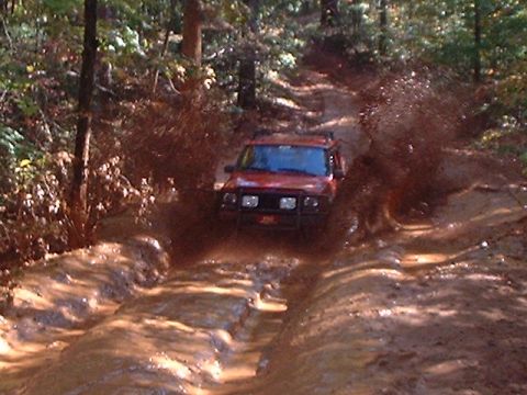 1990 Ford Bronco - Red SUV navigating a muddy off-road trail with significant water spray.