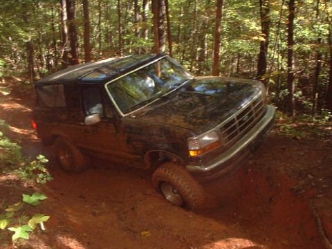 1990 Ford Bronco - A muddy black Ford Bronco navigates a wooded trail.