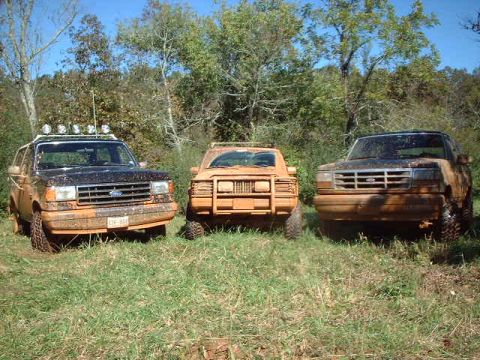 1990 Ford Bronco - Three Ford Broncos are parked in a grassy, wooded area, covered in mud.
