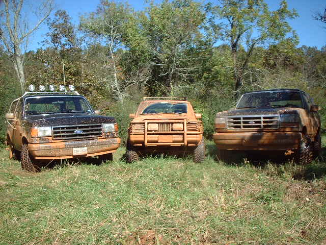 Three muddy lifted Ford trucks parked in a grassy field, front view
